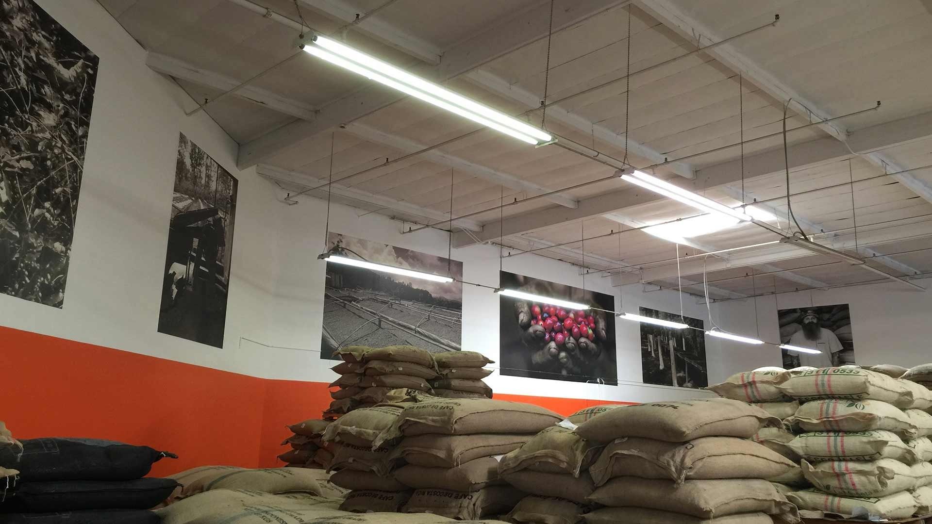 Large stacks of burlap coffee sacks fill a warehouse with white walls, an orange lower section, and hanging photo posters.