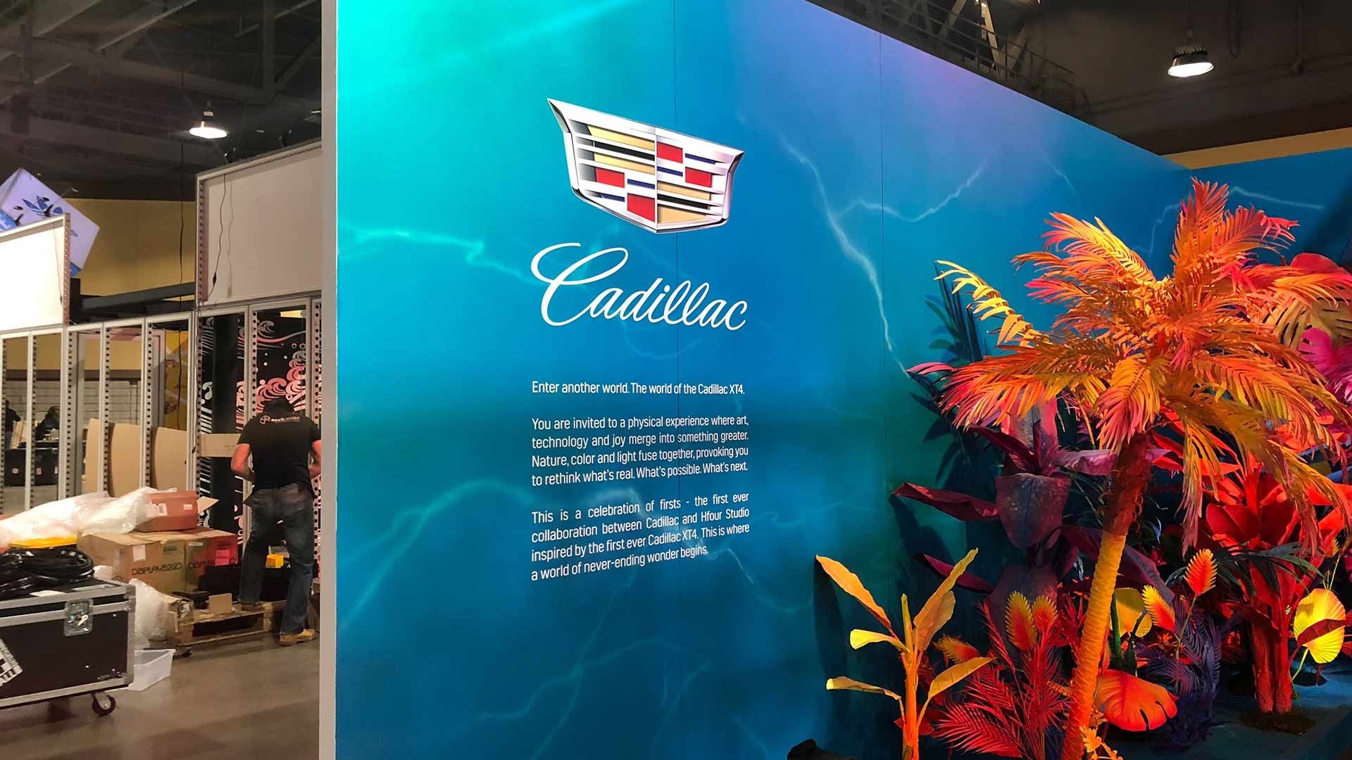 A Cadillac branded sign against a blue background with colorful, stylized tropical plants at a trade show exhibit.