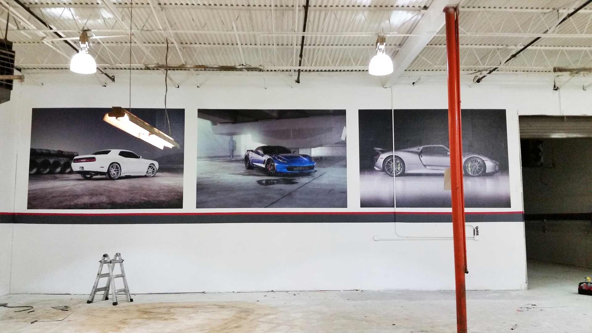 Three large wall posters of sports cars displayed in an industrial space with a ladder and red structural pole.