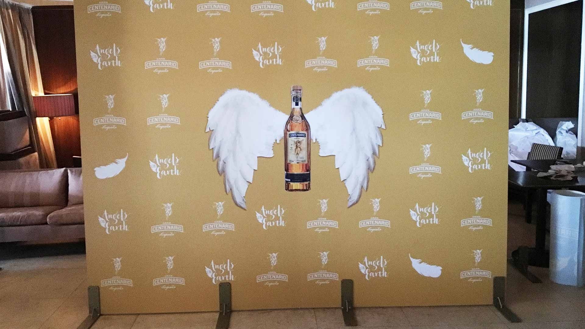 A gold step-and-repeat backdrop featuring a bottle of Black Angel tequila with large white wings at its center.
