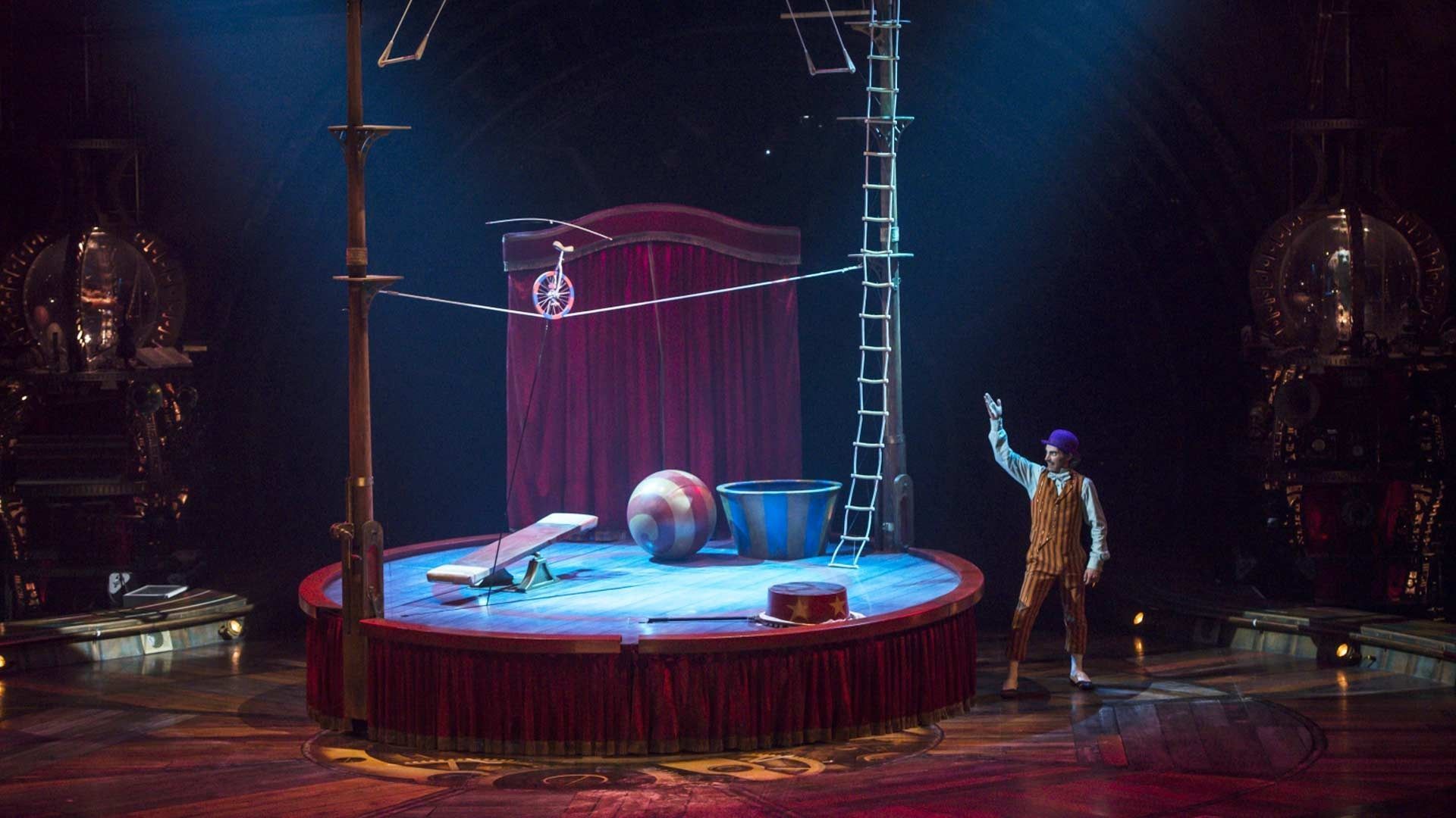 A performer in a brown suit stands on a circular circus stage with a tightrope, ladder, and props under blue spotlights.