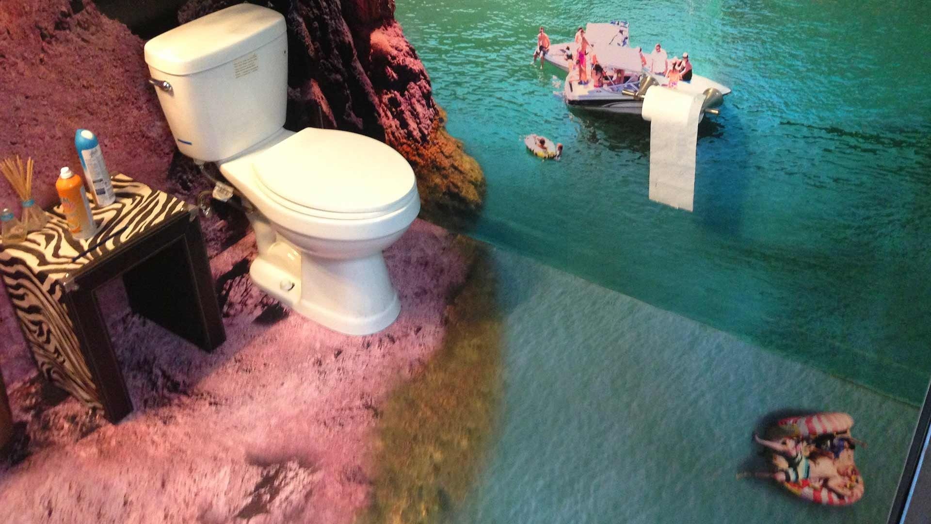 A toilet sits next to a side table, with a 3D floor mural creating an optical illusion of a beach and ocean.