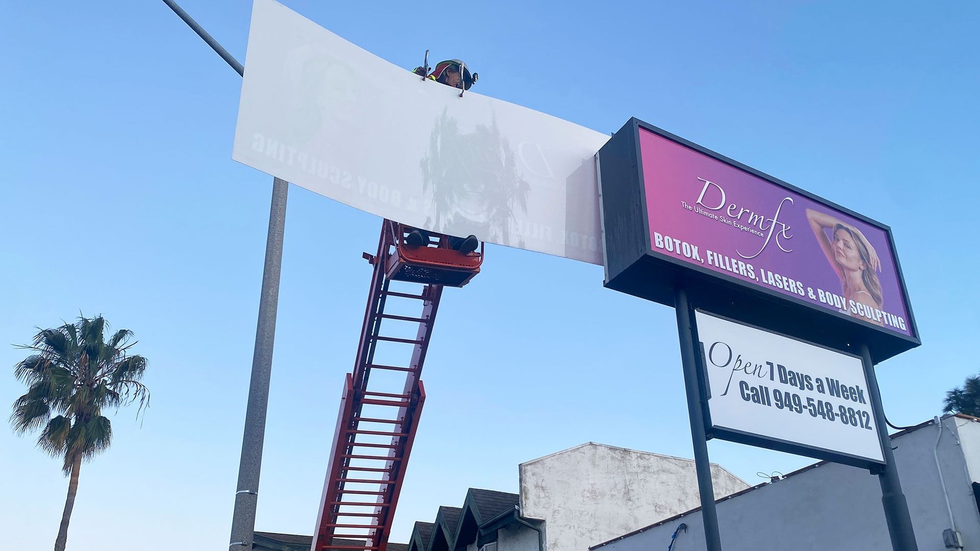 A worker in a cherry picker bucket installs a blank white sign next to a purple and black retail business sign.
