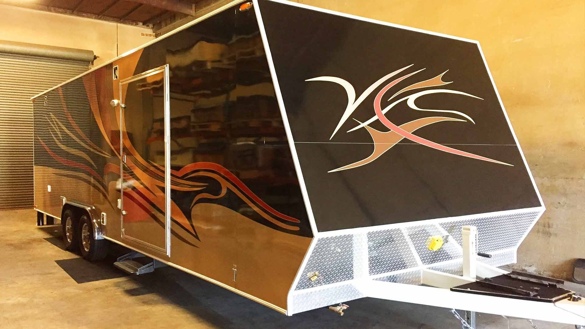 A large black and bronze car trailer with custom tribal-style graphics, parked inside a garage.