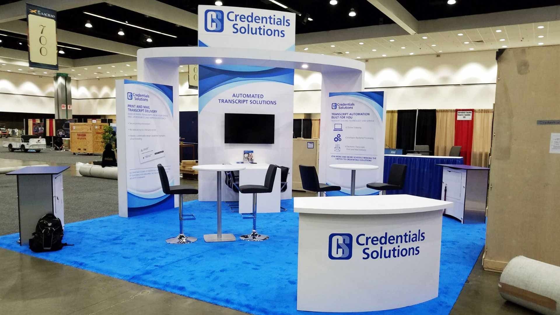 A trade show booth with blue carpeting, white counters, bar stools, and signage for 