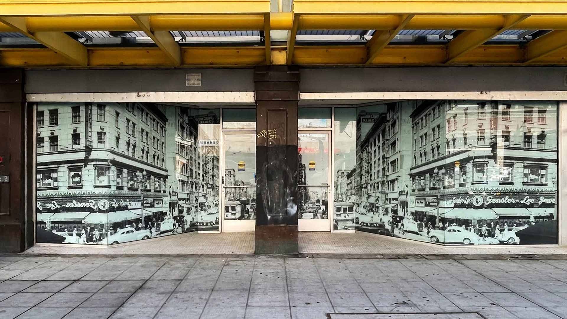 A storefront with windows covered in a black-and-white mural of a vintage city street under a yellow architectural frame.