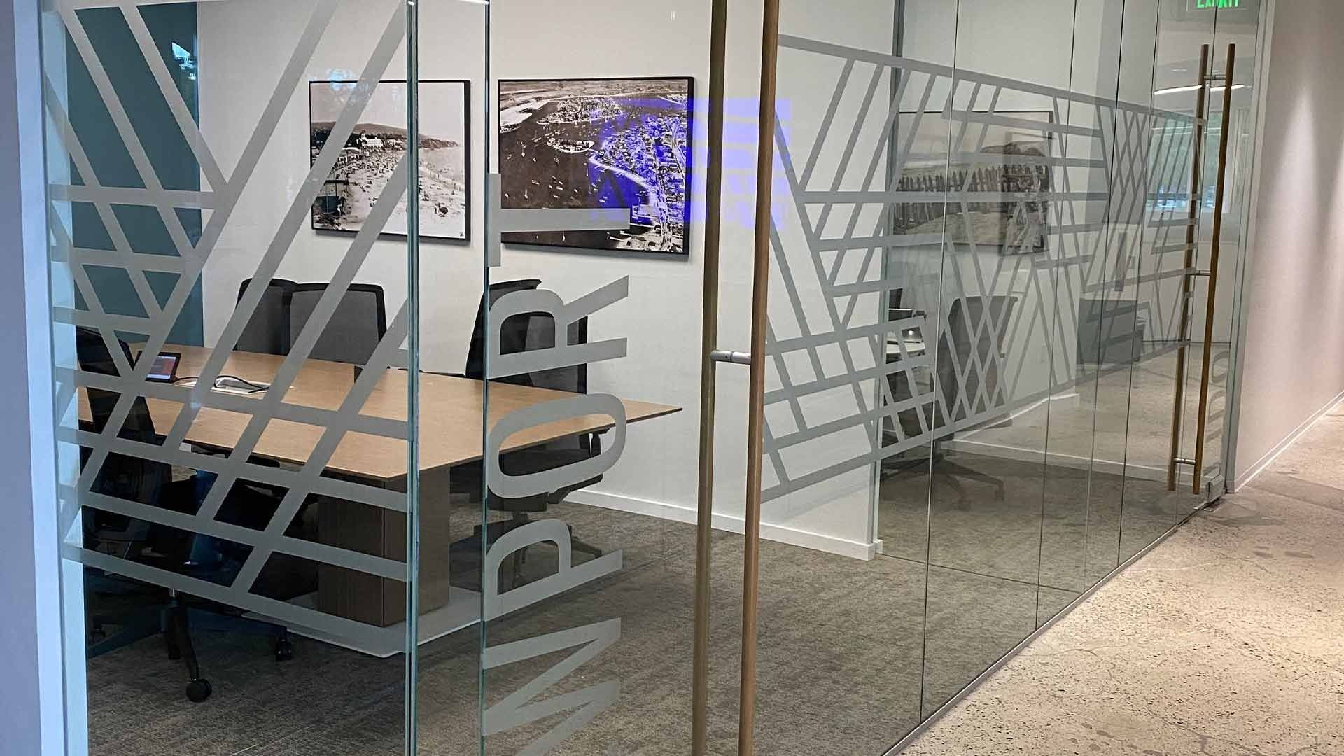 A glass-walled office conference room with a wooden table, chairs, and modern patterned decals on the glass partitions.