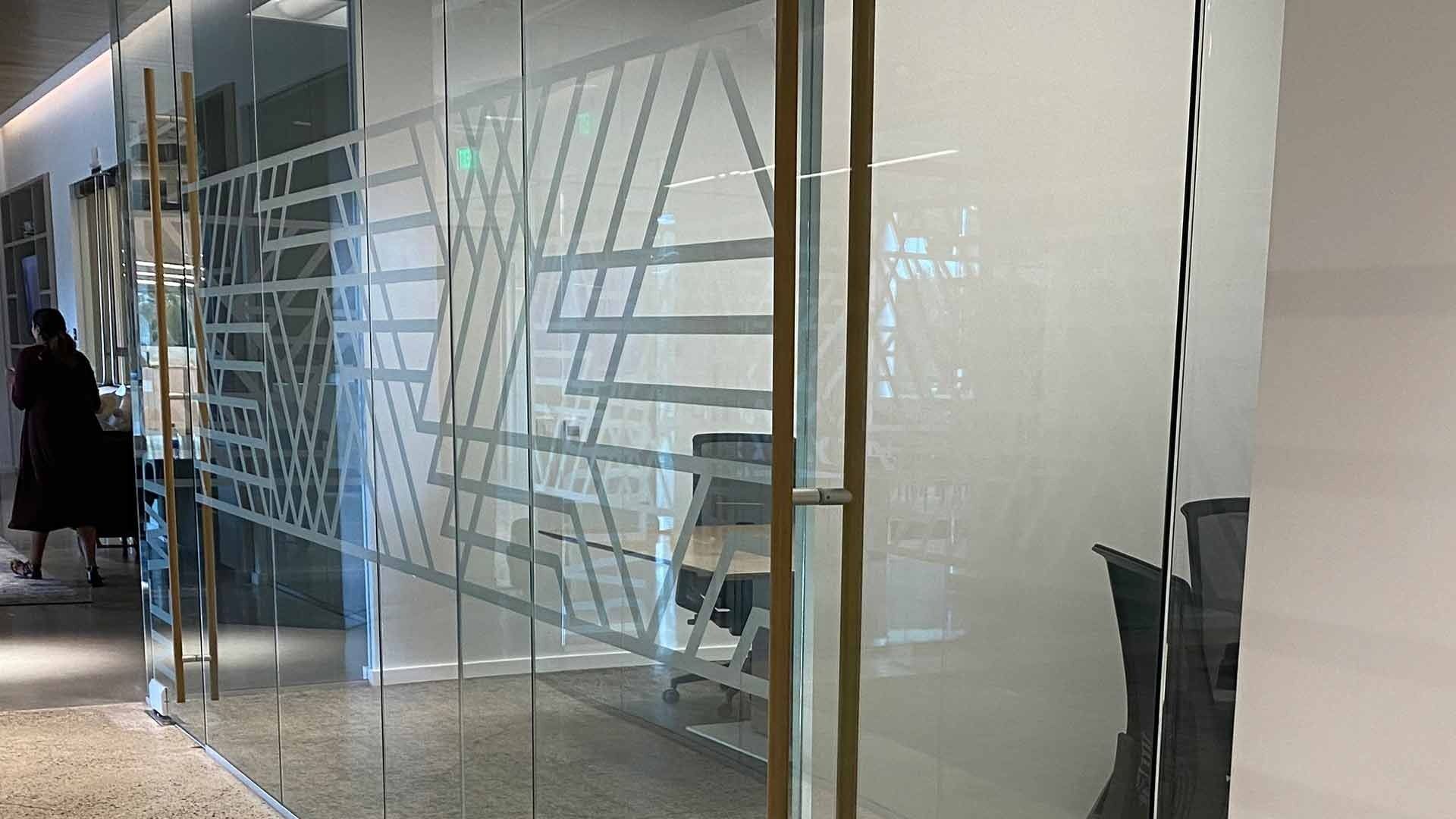 A glass office partition featuring a frosted geometric pattern, with a person walking past in a blurred hallway.