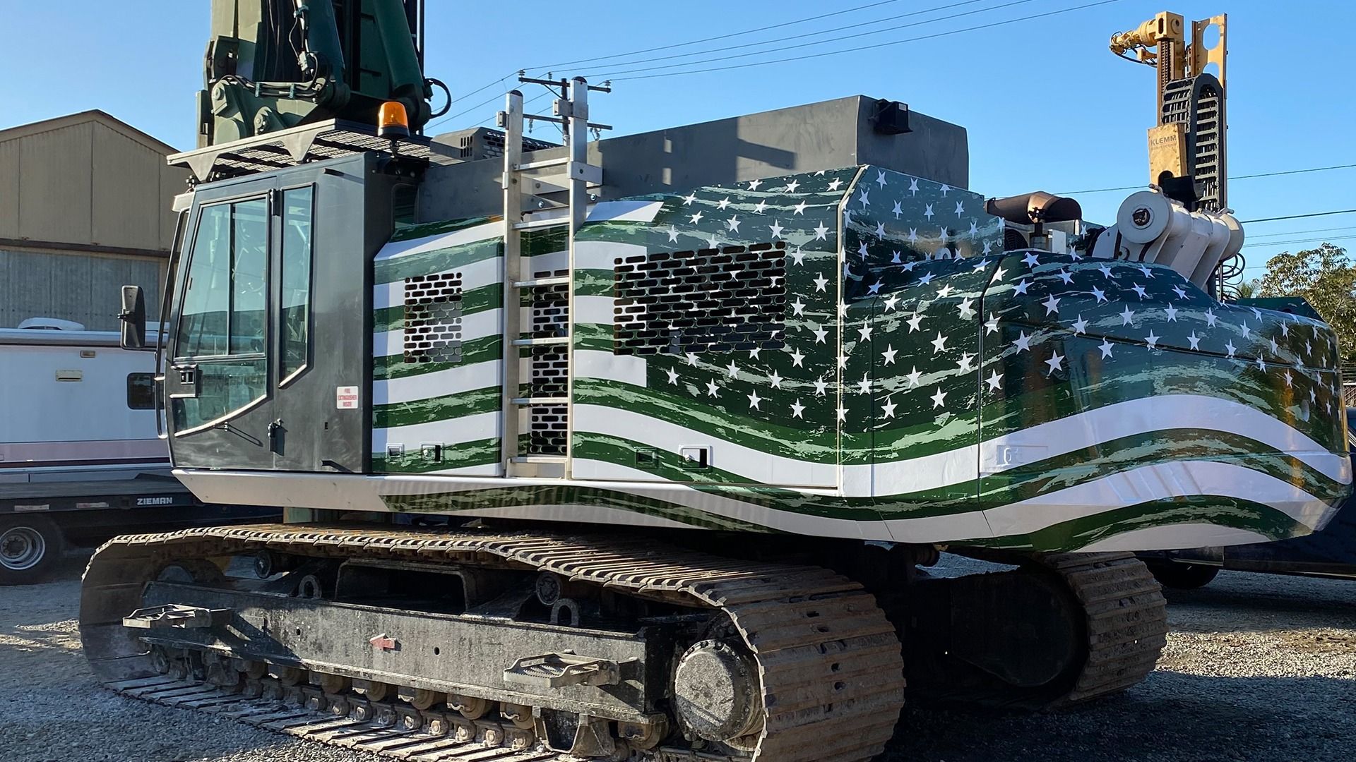 A heavy machinery crawler unit with a green and white American flag wrap, parked on a gravel lot under a clear sky.