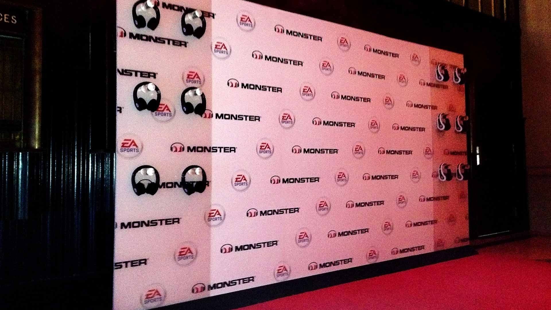 A pink step-and-repeat wall featuring Monster and EA logos with headphones mounted on the surface above a red carpet.