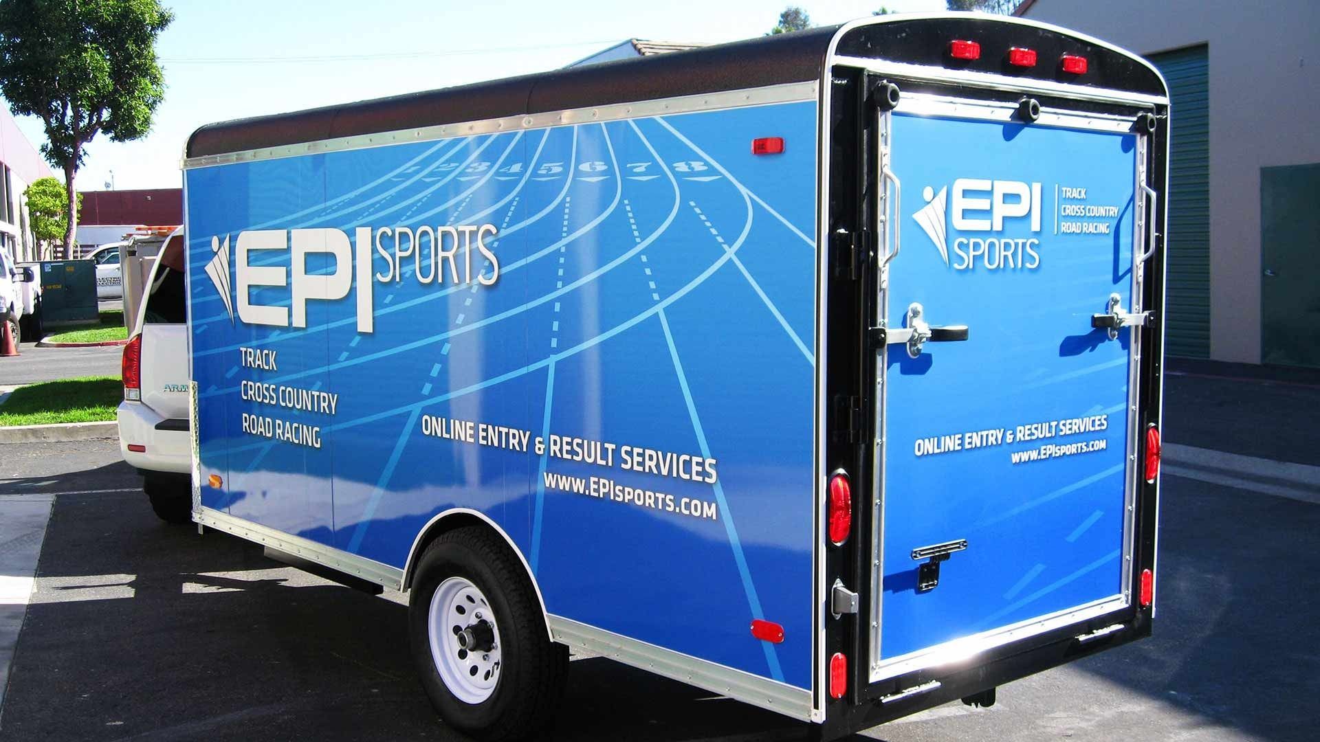 A blue, branded EPI Sports cargo trailer parked outdoors, featuring a race track graphic on its side and back doors.