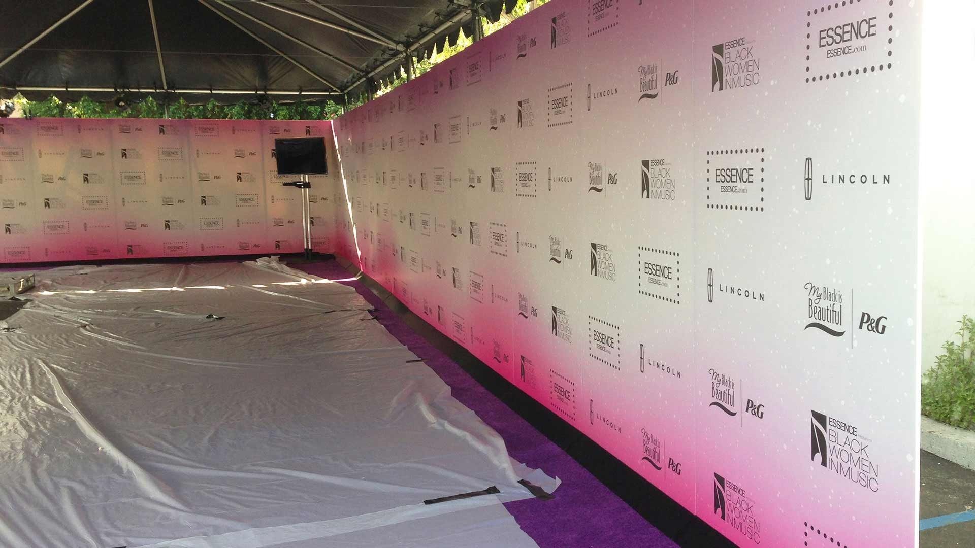 An L-shaped media wall with a pink gradient background features repeated sponsor logos under a tent at an event.