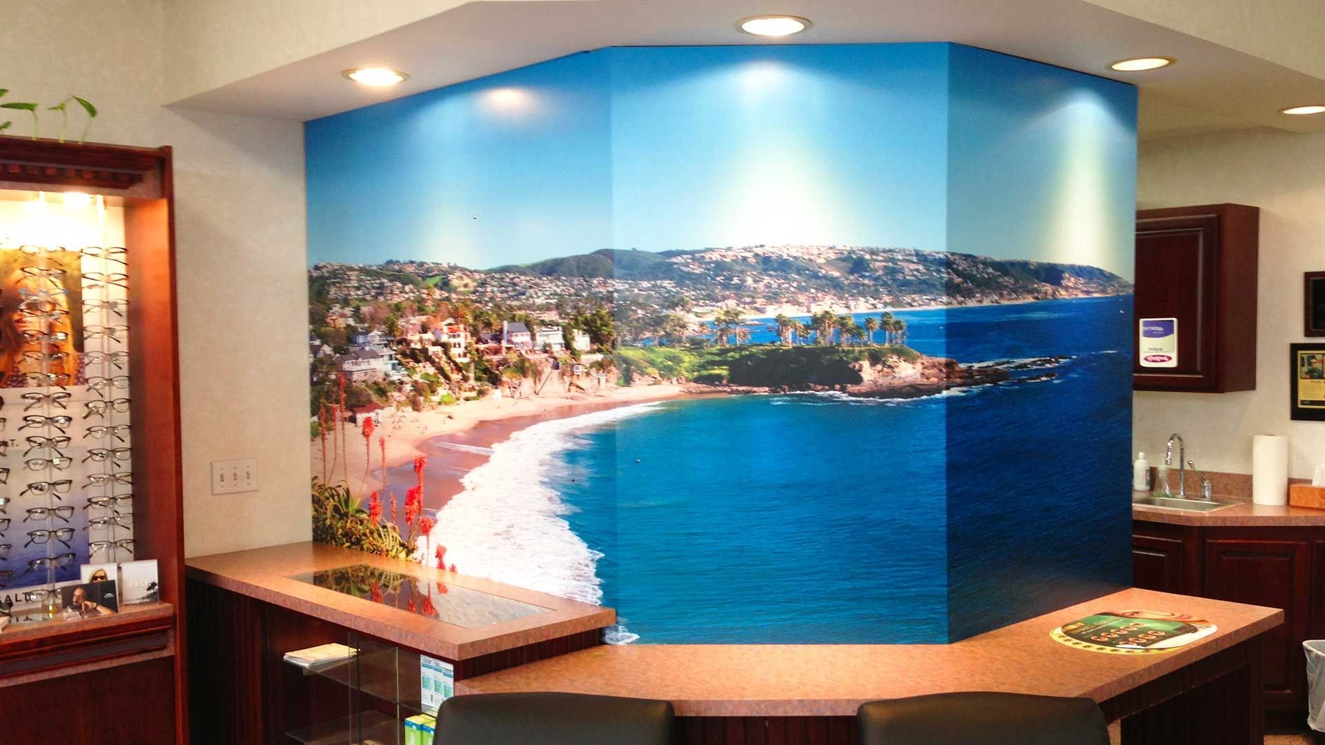 A large panoramic ocean beach mural spans a multi-panel office wall behind a wooden reception counter.
