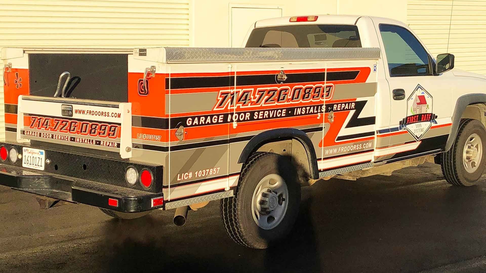 A white service truck with orange and black graphics for a garage door repair company parked on a paved lot.