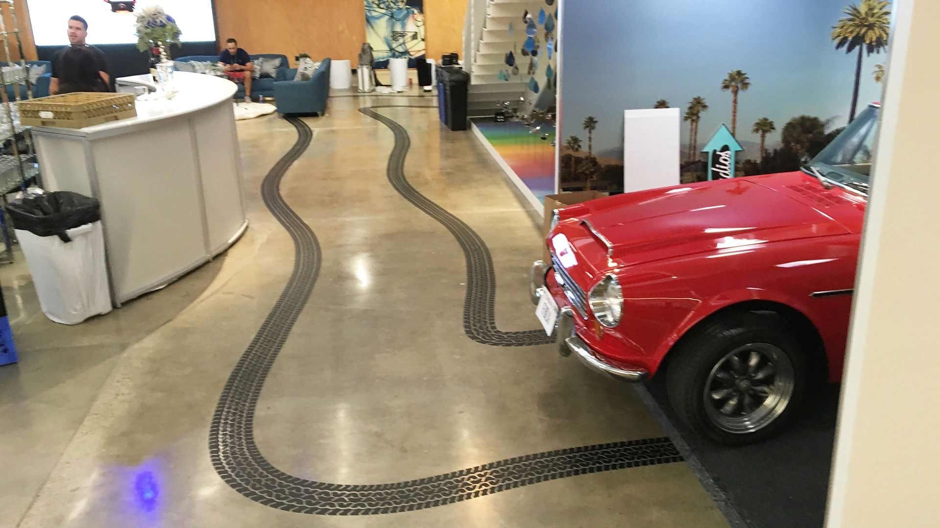 A red vintage convertible sits on display with decorative tire tracks painted on the floor leading toward it.