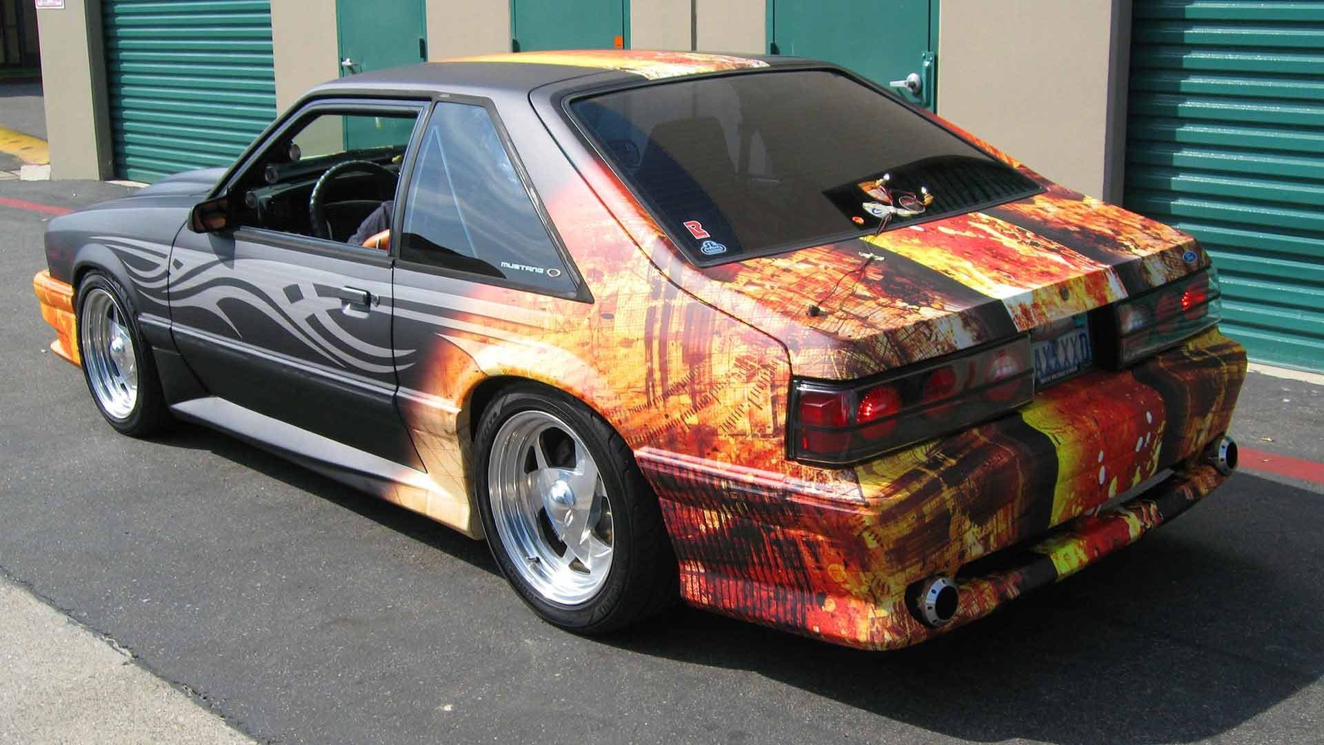A modified Fox-body Ford Mustang with a custom fiery orange and yellow wrap parked outside a storage facility.