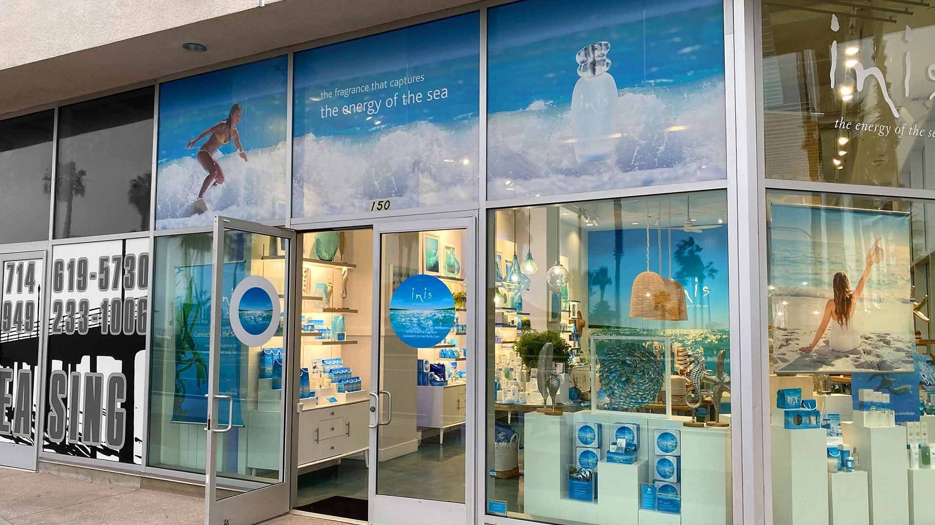 A storefront with blue-tinted windows featuring beach imagery, interior product displays, and a leasing sign on the left.