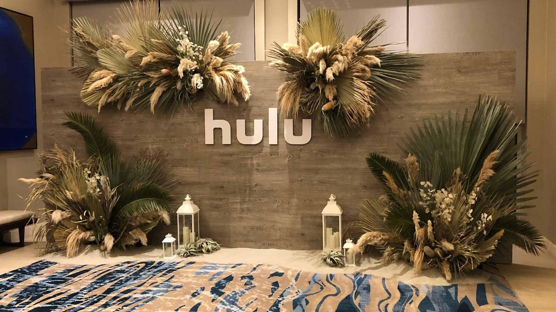 A Hulu-branded event backdrop with a wood-paneled wall, dried floral arrangements, lanterns, and a blue patterned rug.