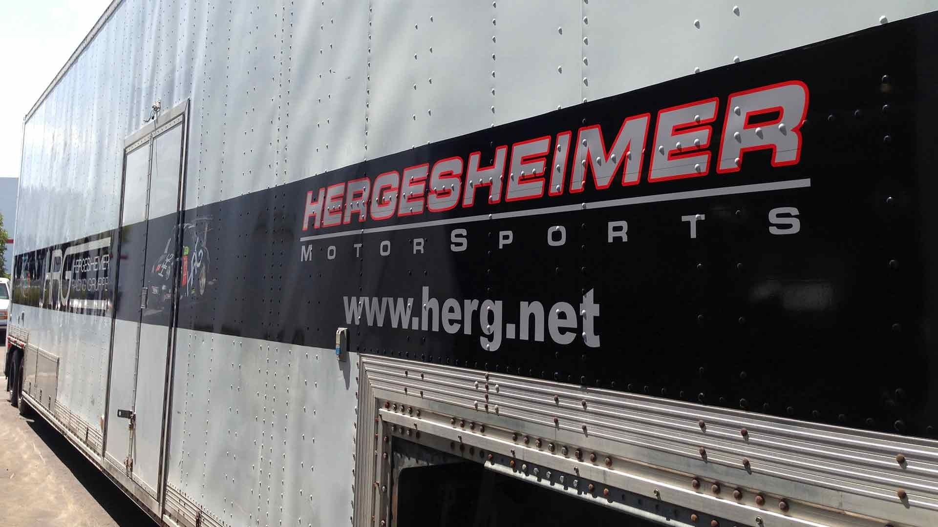 Side view of a large white motorsport trailer with a black stripe featuring the 