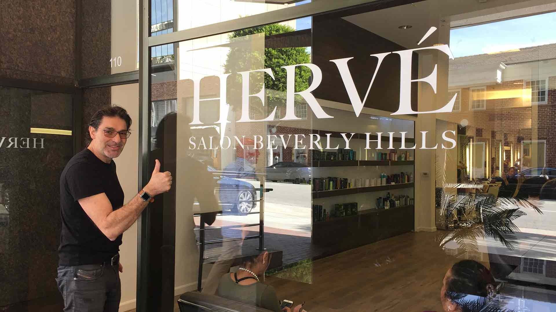 A man in a black shirt gives a thumbs-up outside Hervé Salon Beverly Hills, seen through its glass storefront.
