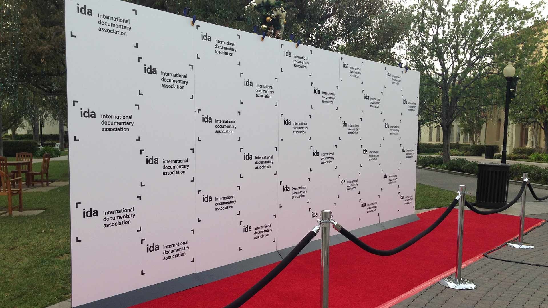 A red carpet leads to a large white step-and-repeat wall featuring repeating IDA logos, set outdoors in a grassy area.