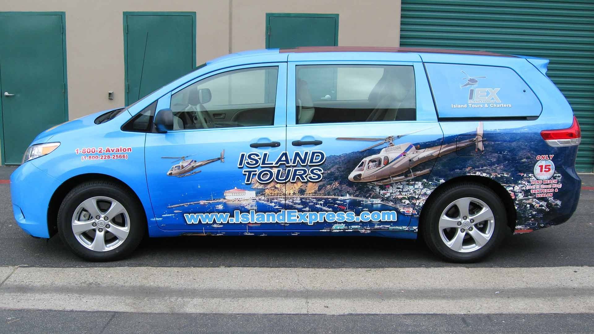 A light blue minivan wrapped in advertisements for Island Tours with helicopter graphics, parked by a building.