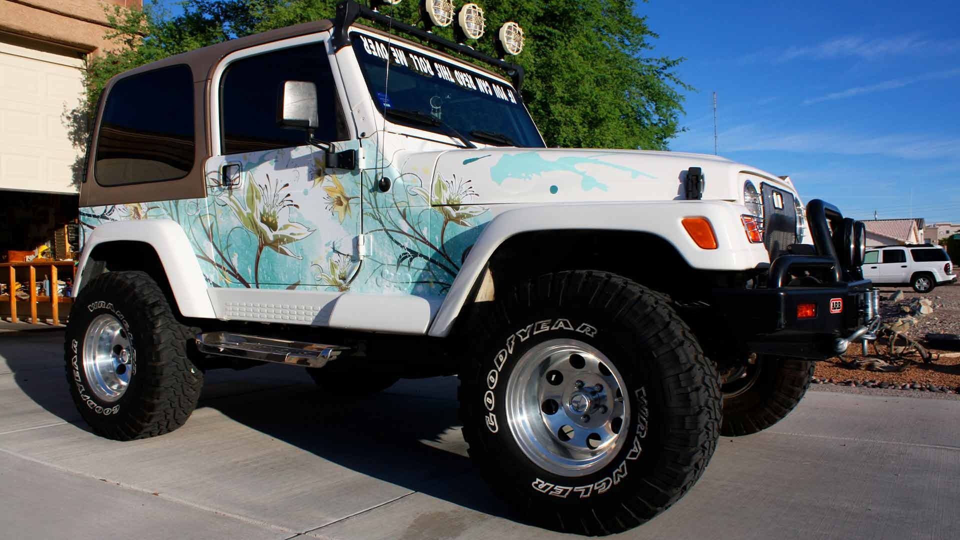 A white Jeep Wrangler with a blue floral wrap, lifted suspension, and large off-road tires parked on a paved driveway.