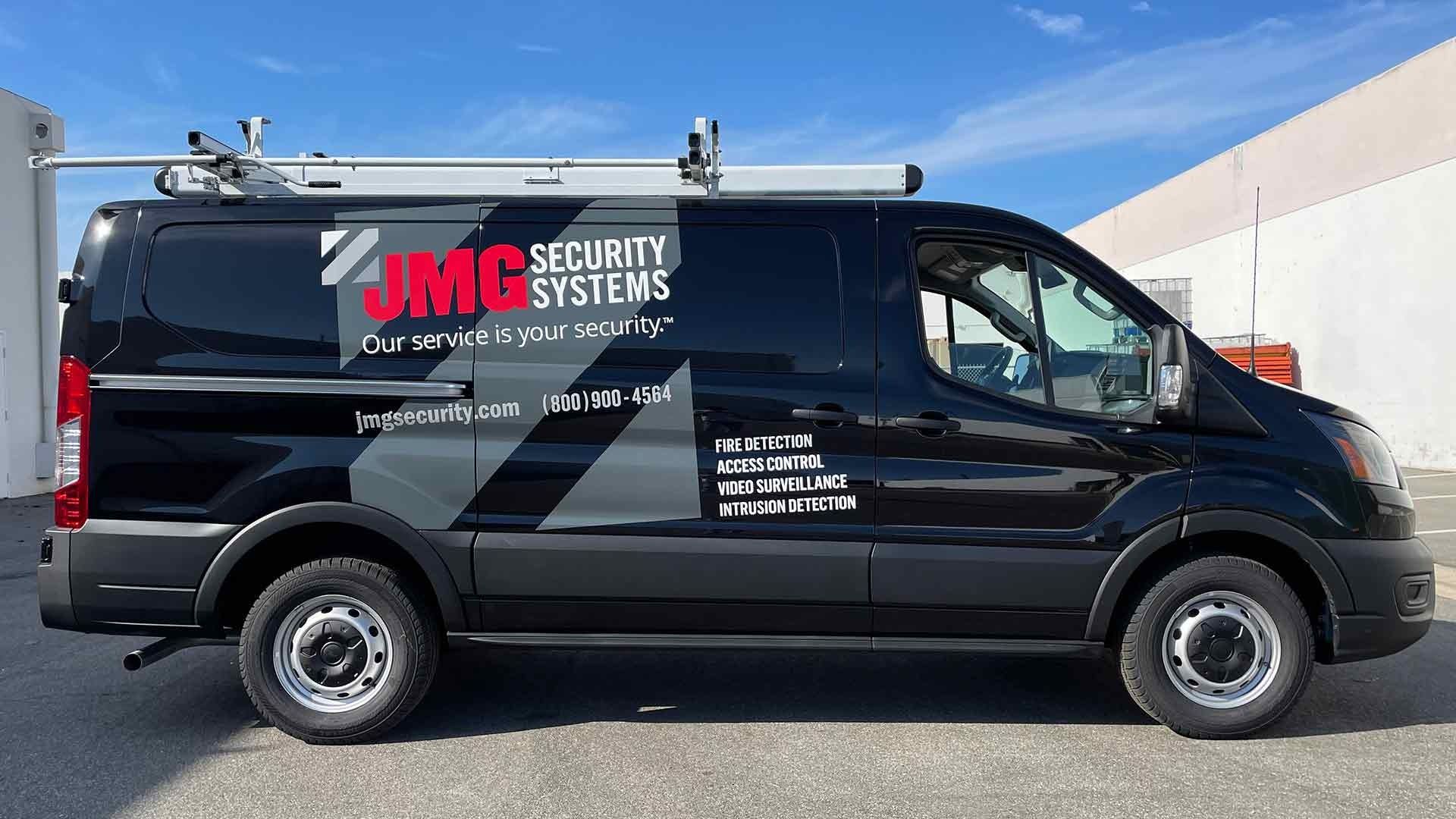 A black JMG Security Systems service van parked in a lot against a bright blue sky.