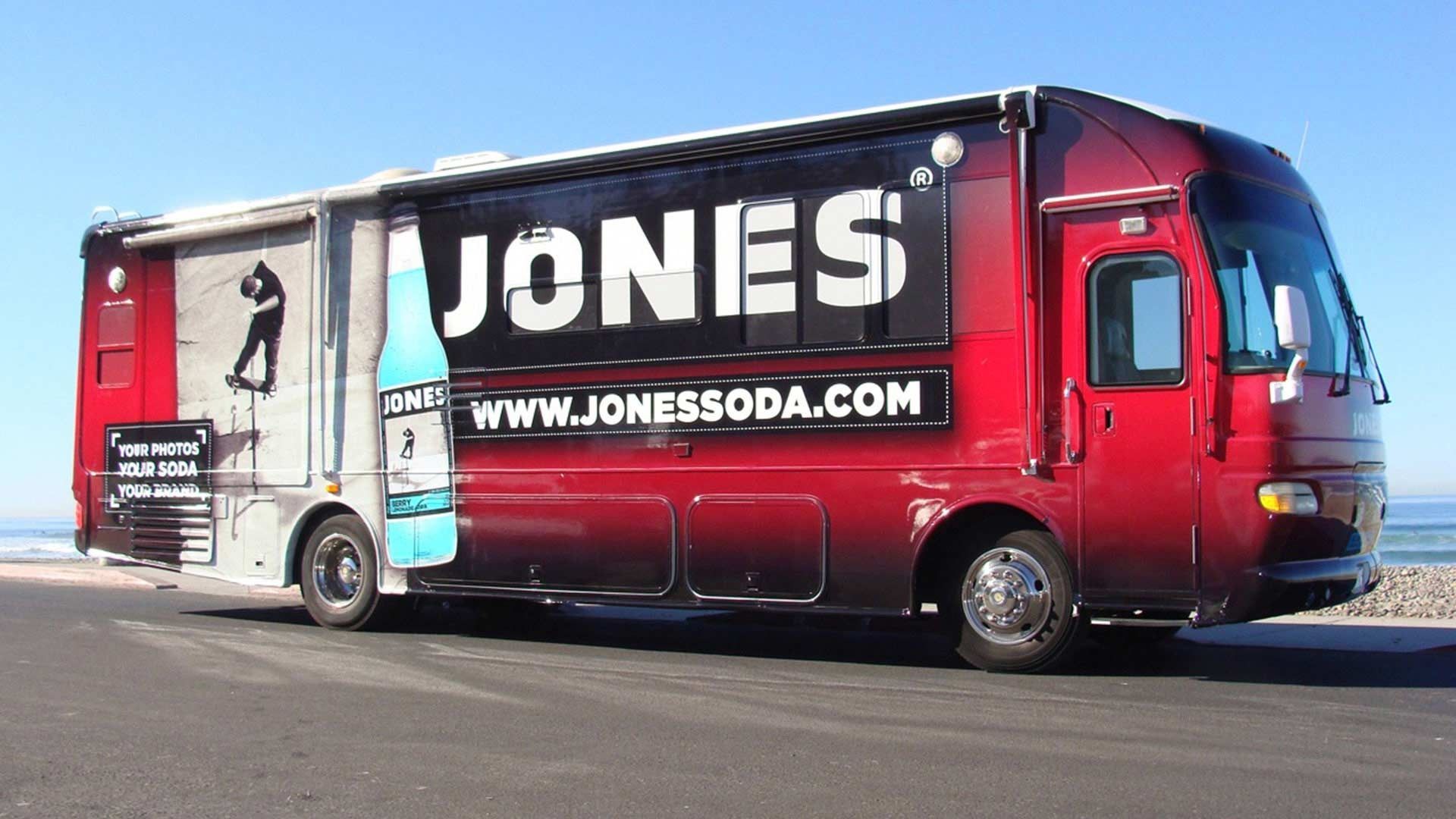 A red promotional bus for Jones Soda parked by the ocean with the brand logo and website displayed on its side.