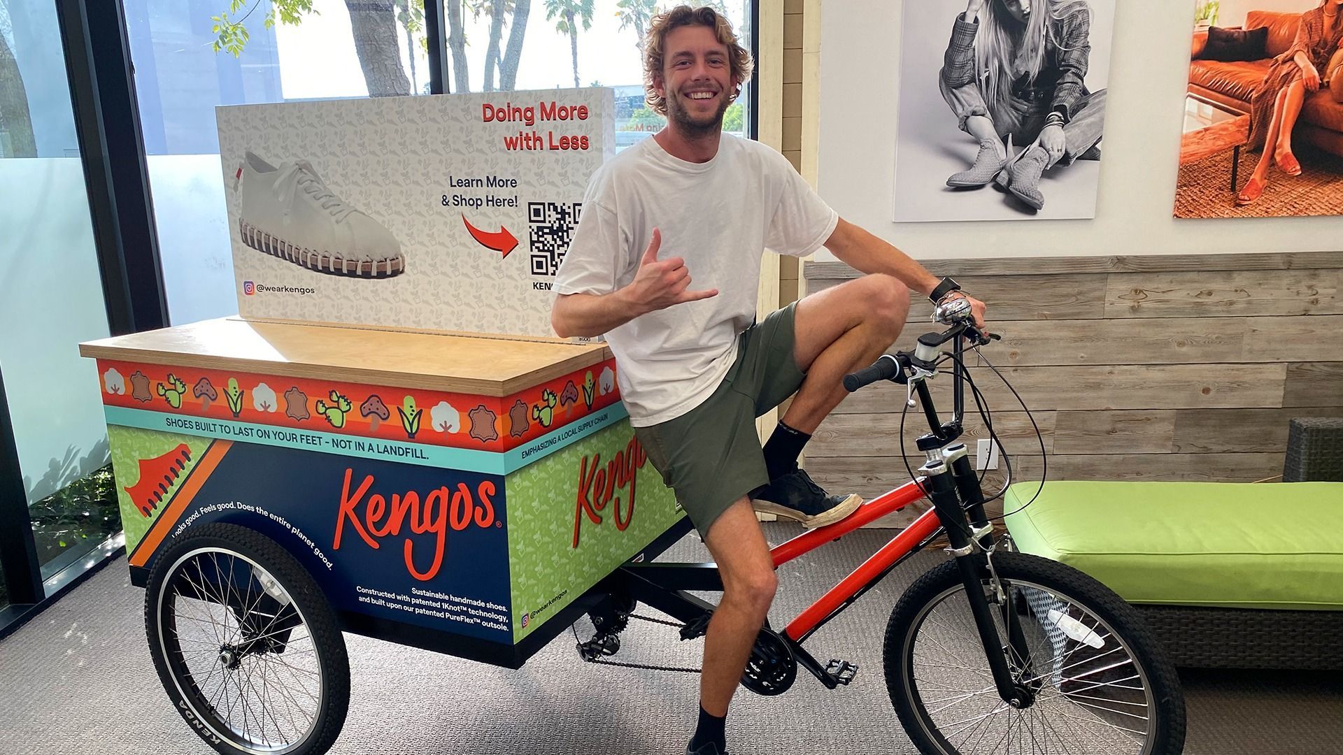A smiling person sits on a branded Kengos cargo tricycle inside an office, making a shaka sign with their hand.
