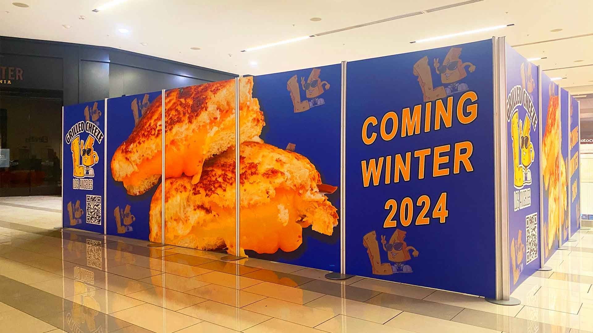 A blue construction wall in a mall featuring images of grilled cheese sandwiches and text reading 