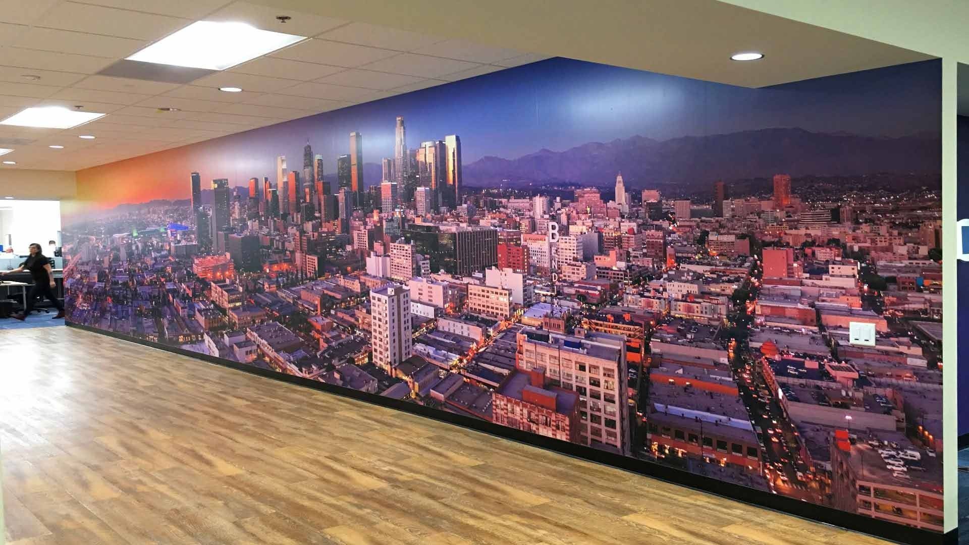 A wide wall mural depicting a panoramic aerial view of the Los Angeles skyline at dusk inside a modern office space.