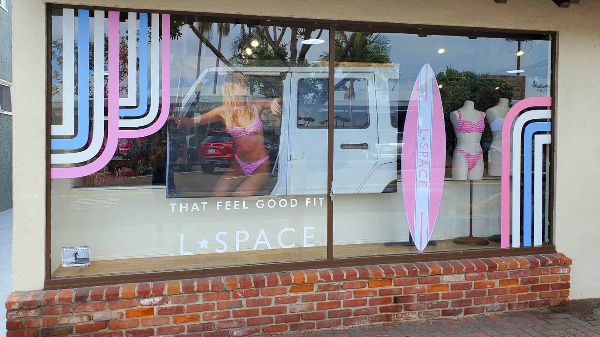 Storefront window display for L*SPACE featuring pink and blue stripes, a surfboard, and a mannequin wearing swimwear.