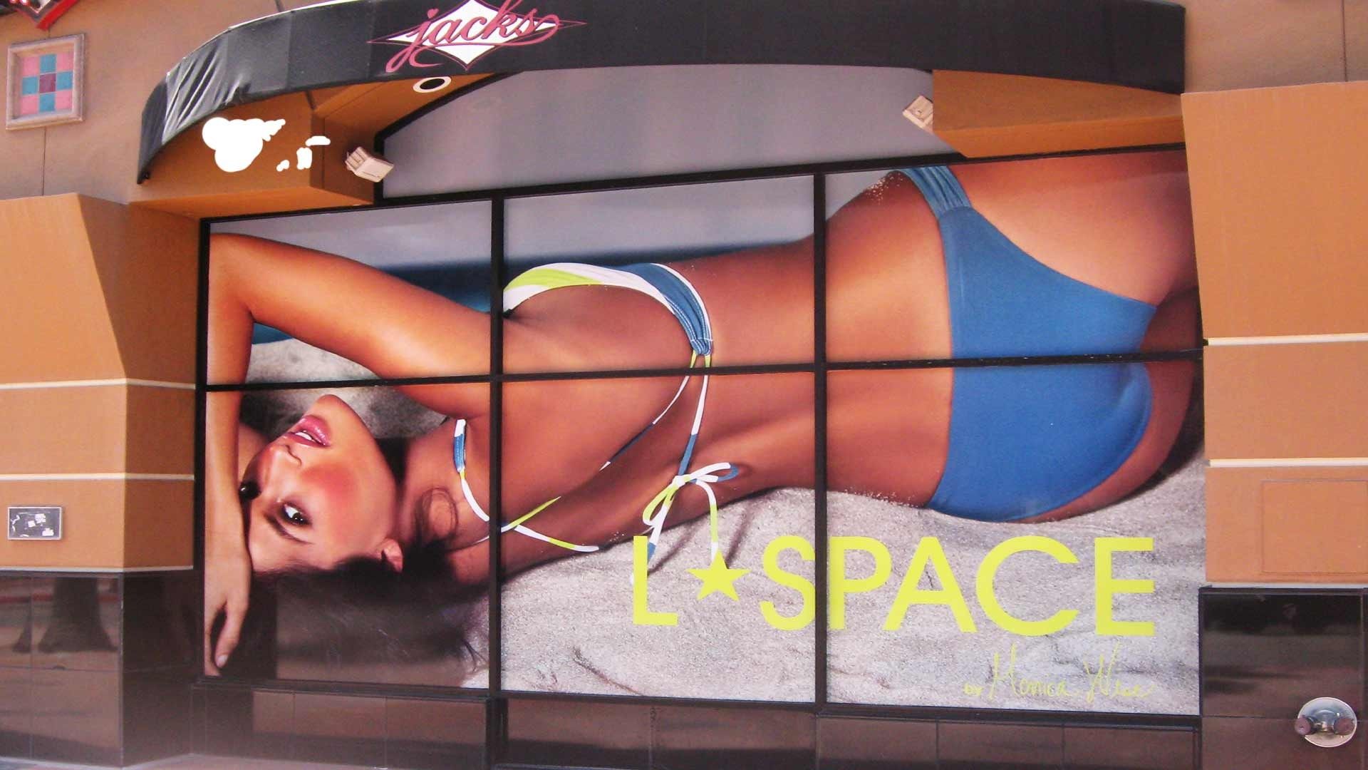 A store window display featuring a large advertisement for L*Space swimwear, showing a person lying on sand.