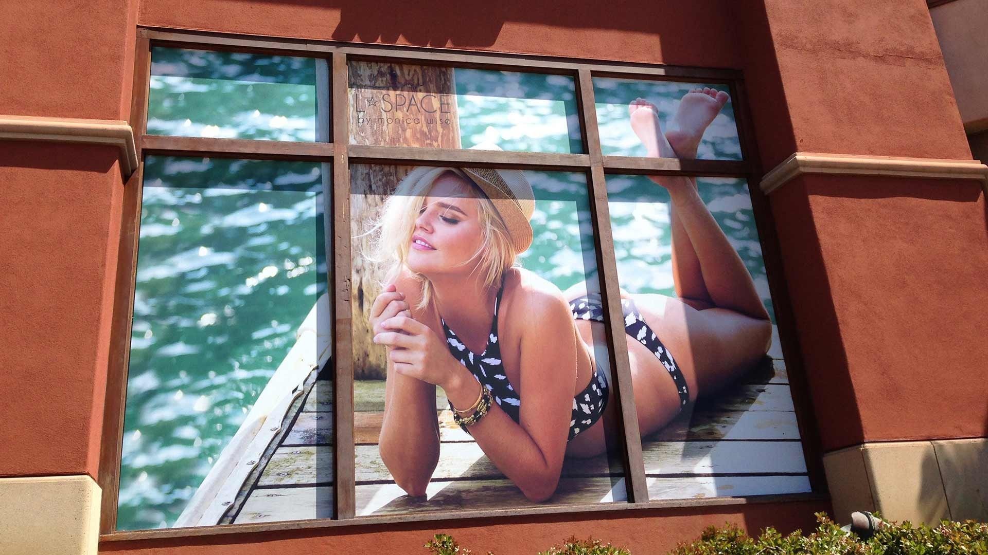 A large window display on a brick building showing a person in a patterned swimsuit lounging on a wooden dock by water.