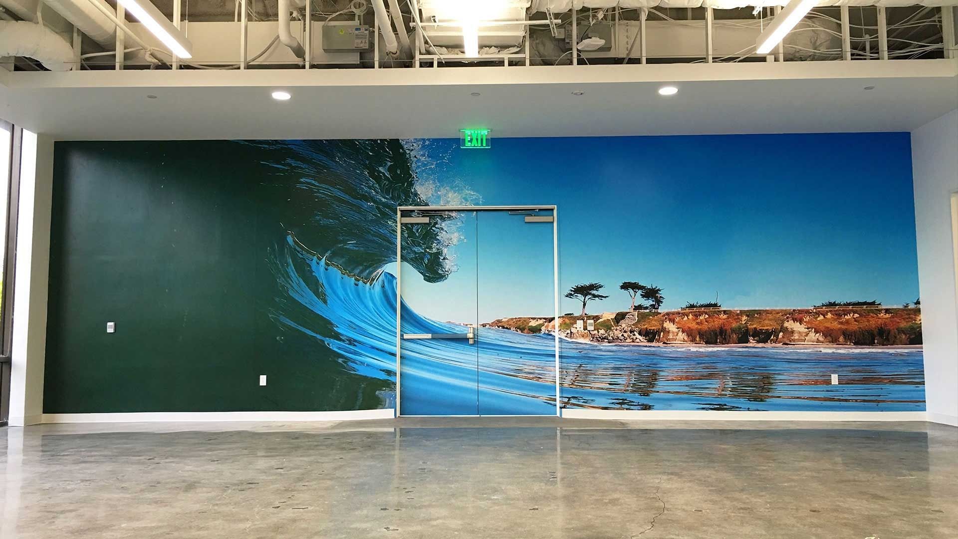 Large wall mural of a crashing ocean wave transitioning into a sunny beach scene, surrounding a set of glass double doors.