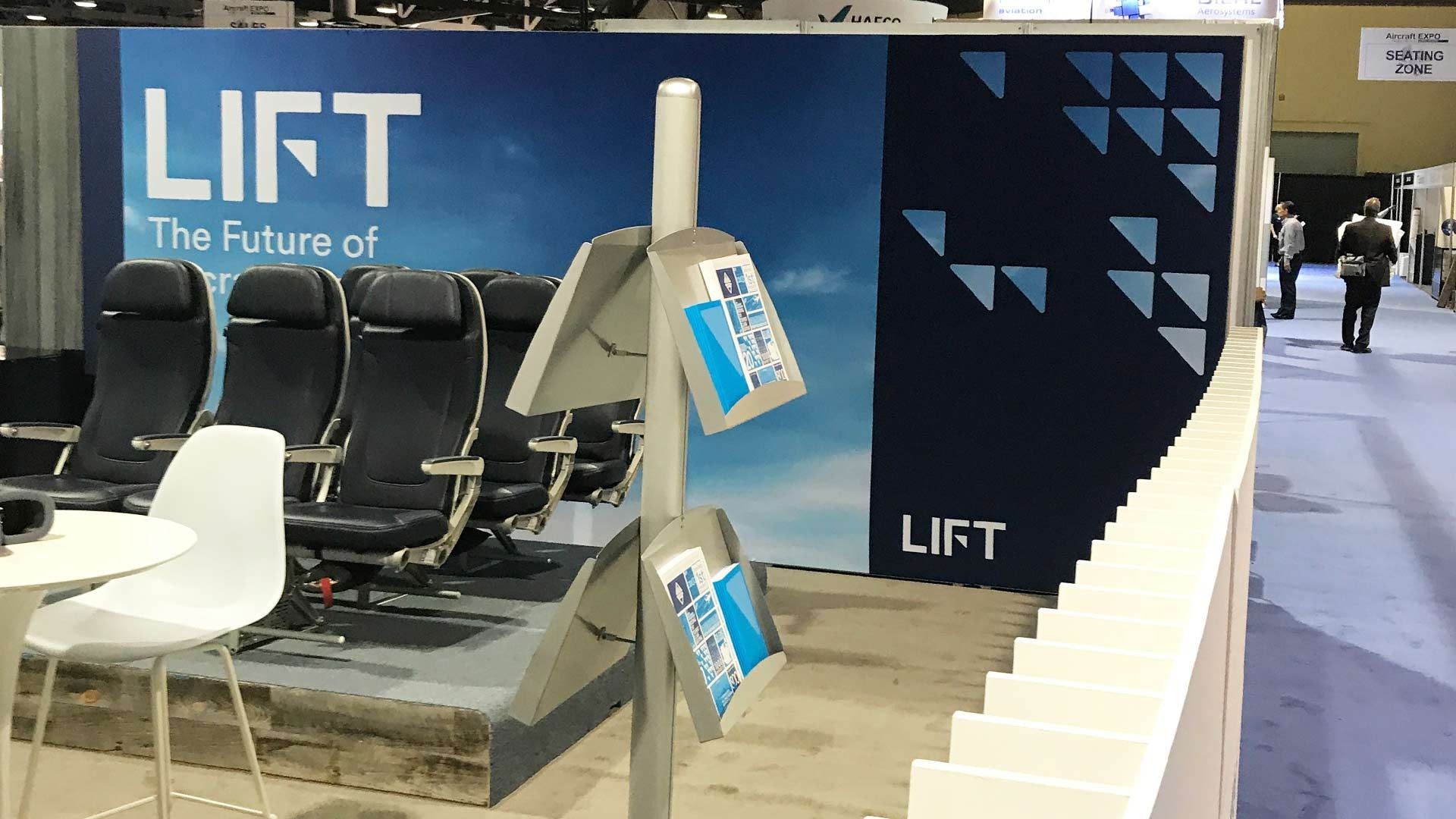A trade show booth for LIFT featuring airline seating on a raised platform with a literature stand in the foreground.