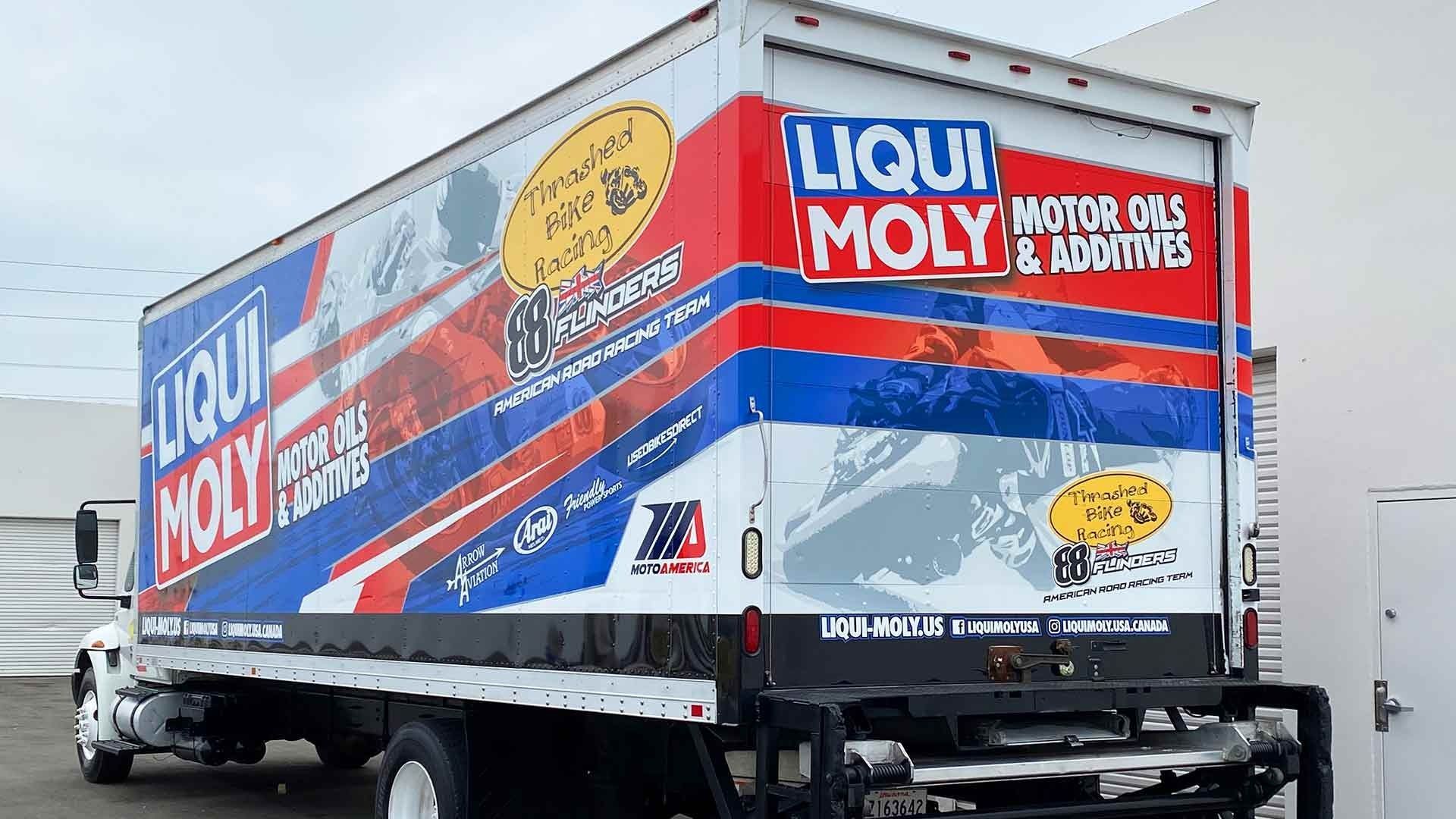 A box truck parked in front of a white building, featuring blue and red Liqui Moly branding on its side and back panels.