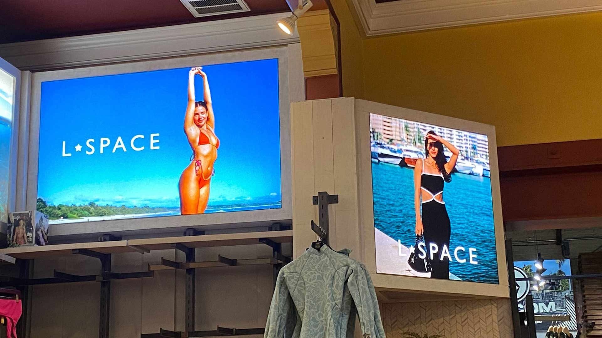 Two illuminated advertising displays for the swimwear brand L*Space, showing models in coastal settings inside a store.