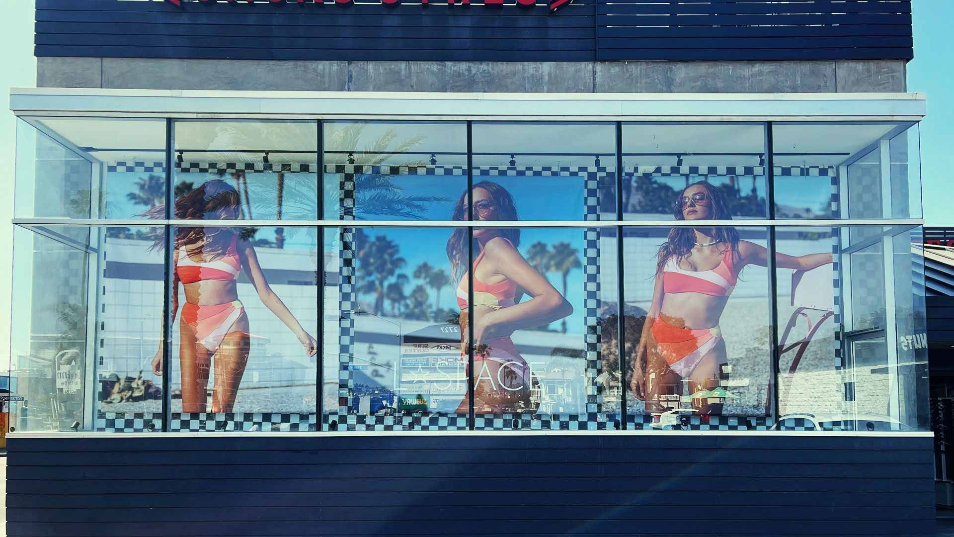 A glass display case shows three panels of a woman in a patterned two-piece swimsuit against a palm tree backdrop.