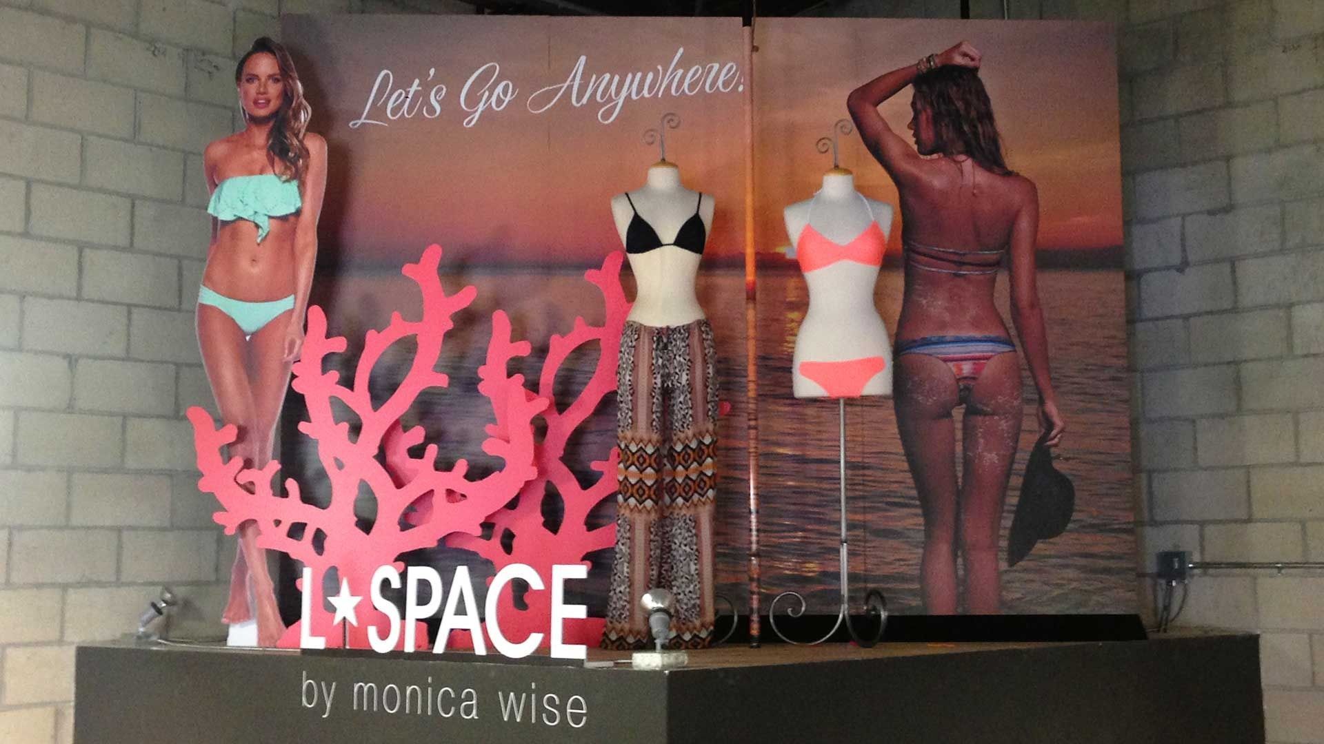 L*Space clothing display featuring bikinis, cover-up pants, and a coral-shaped cutout on a platform against a sunset backdrop.