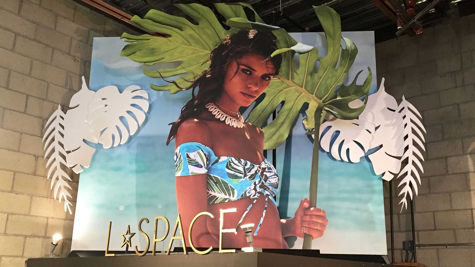 A retail display for L*Space featuring a poster of a person in tropical swimwear, surrounded by large paper palm leaves.