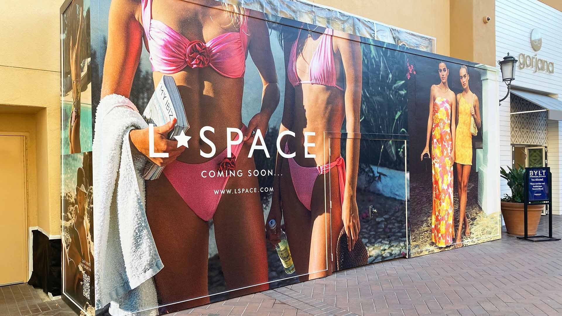 A large, branded construction hoarding for L*SPACE swimwear featuring models in bikinis, advertising a coming soon store.