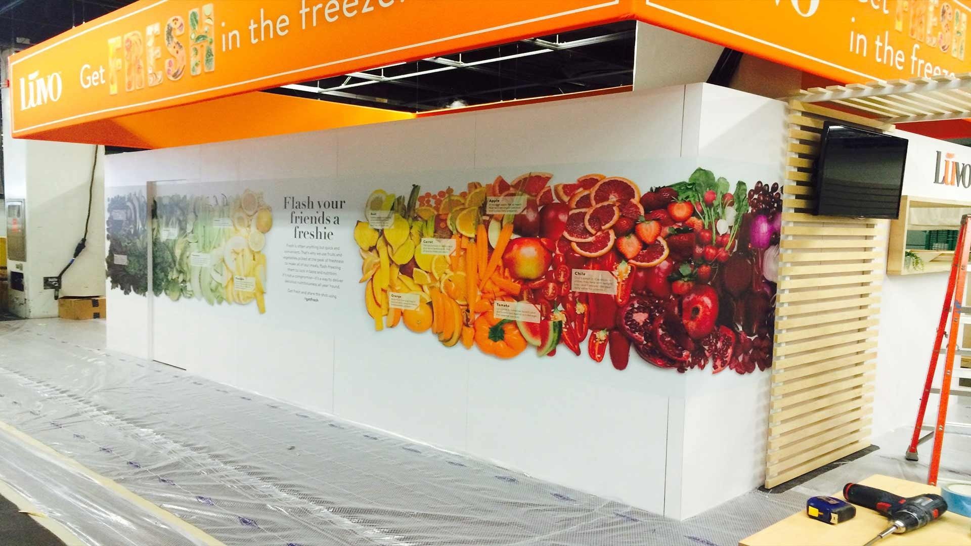 A display booth features a wall mural of a rainbow-colored gradient of fresh produce, topped by an orange overhead sign.