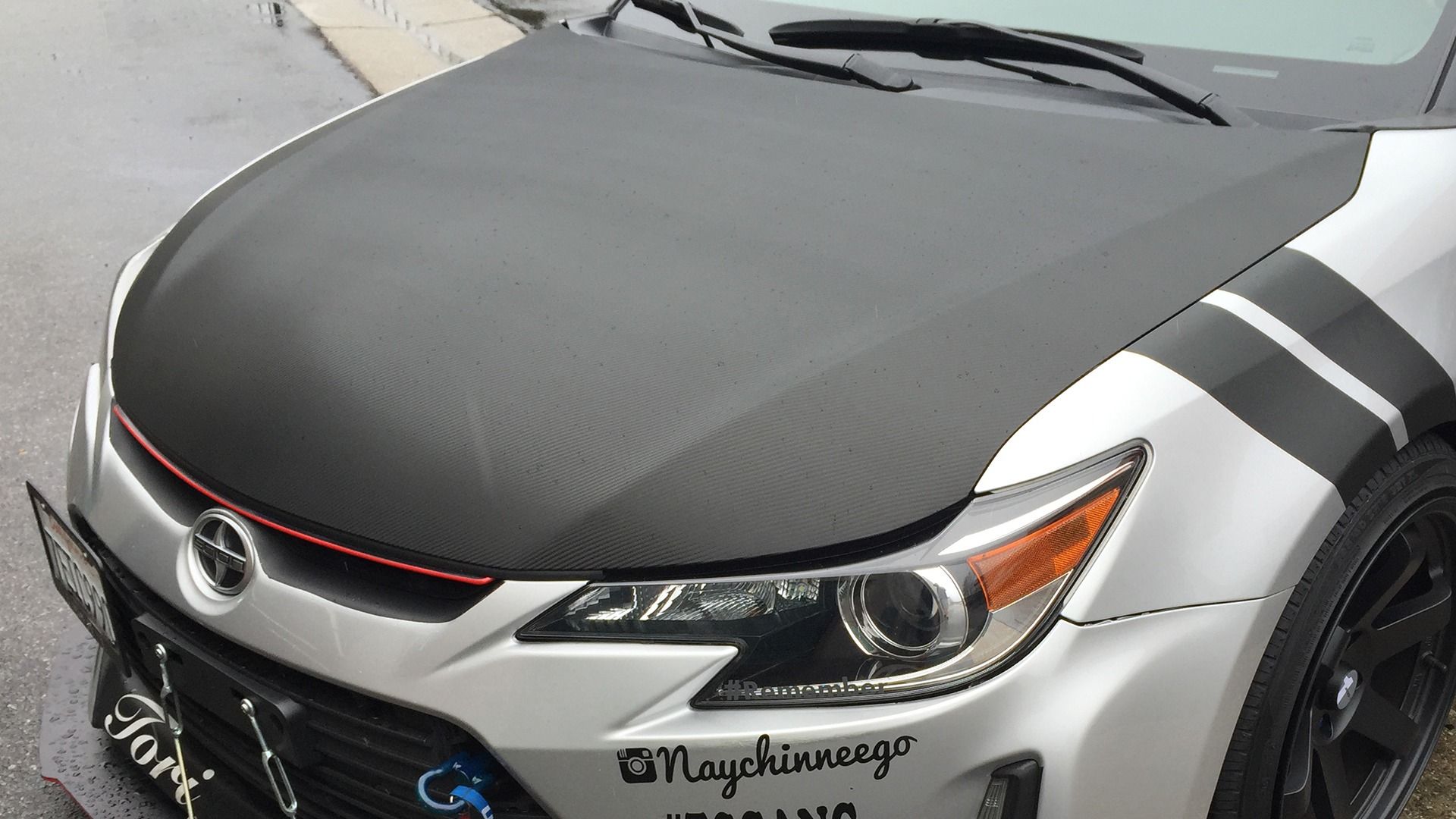 A silver Scion car with a textured black carbon-fiber hood, racing stripes on the fender, and various decals on the bumper.