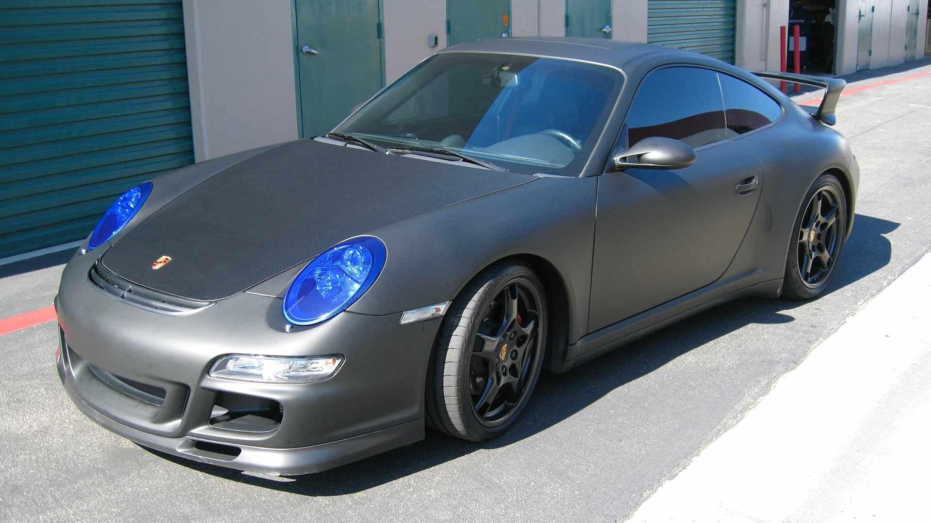 A matte grey Porsche 911 with bright blue headlights parked on pavement in front of storage units.