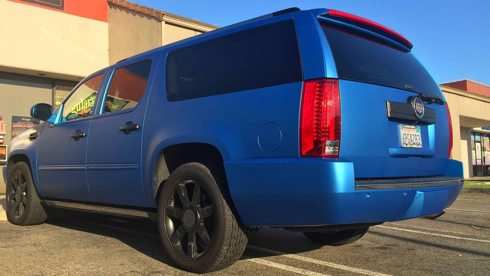 A matte blue Cadillac Escalade SUV with black rims parked in a sunlit parking lot in front of commercial buildings.