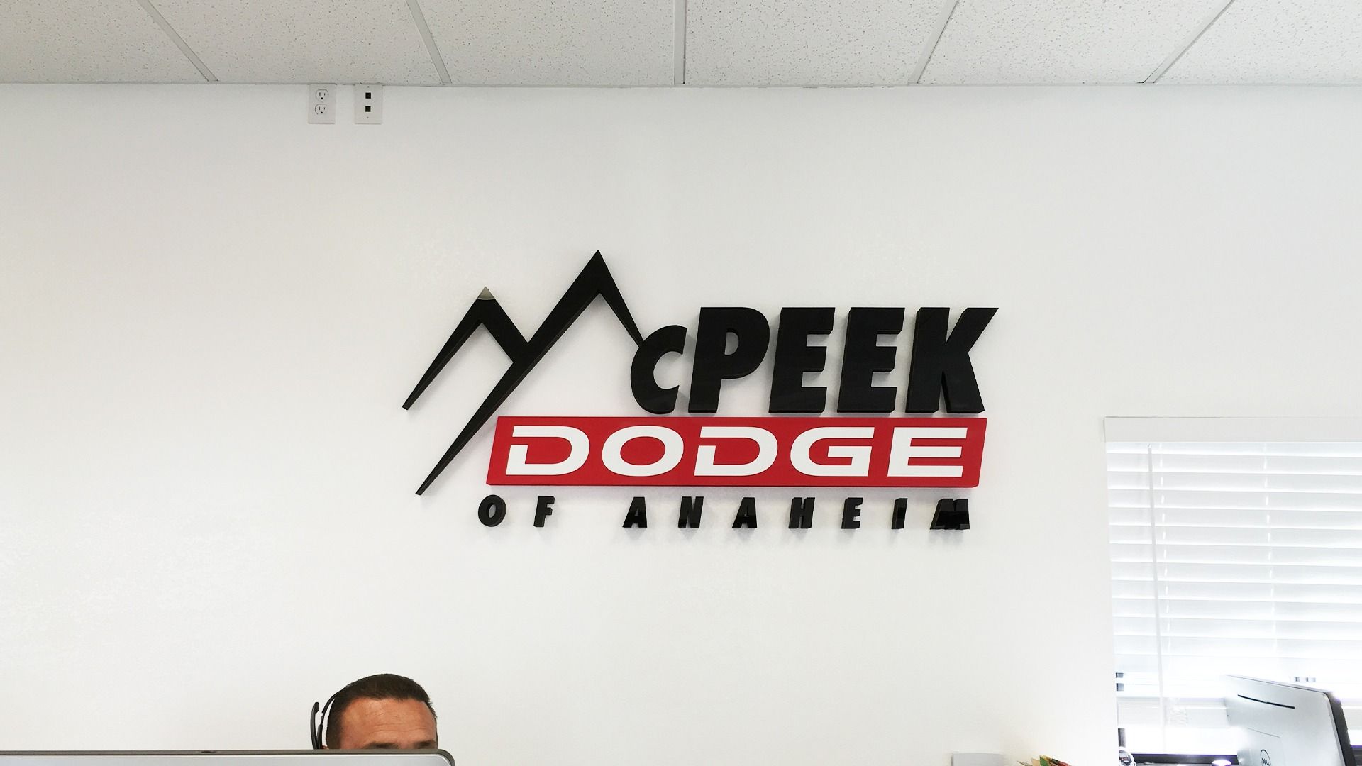 A logo for McPeek Dodge of Anaheim mounted on a white wall above a person working at a desk.