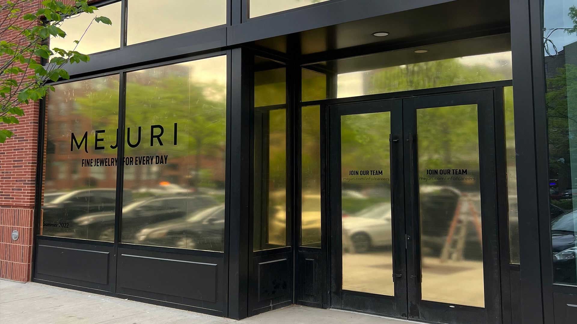 The exterior storefront of a Mejuri jewelry shop featuring black framing and glass windows reflecting cars and trees.