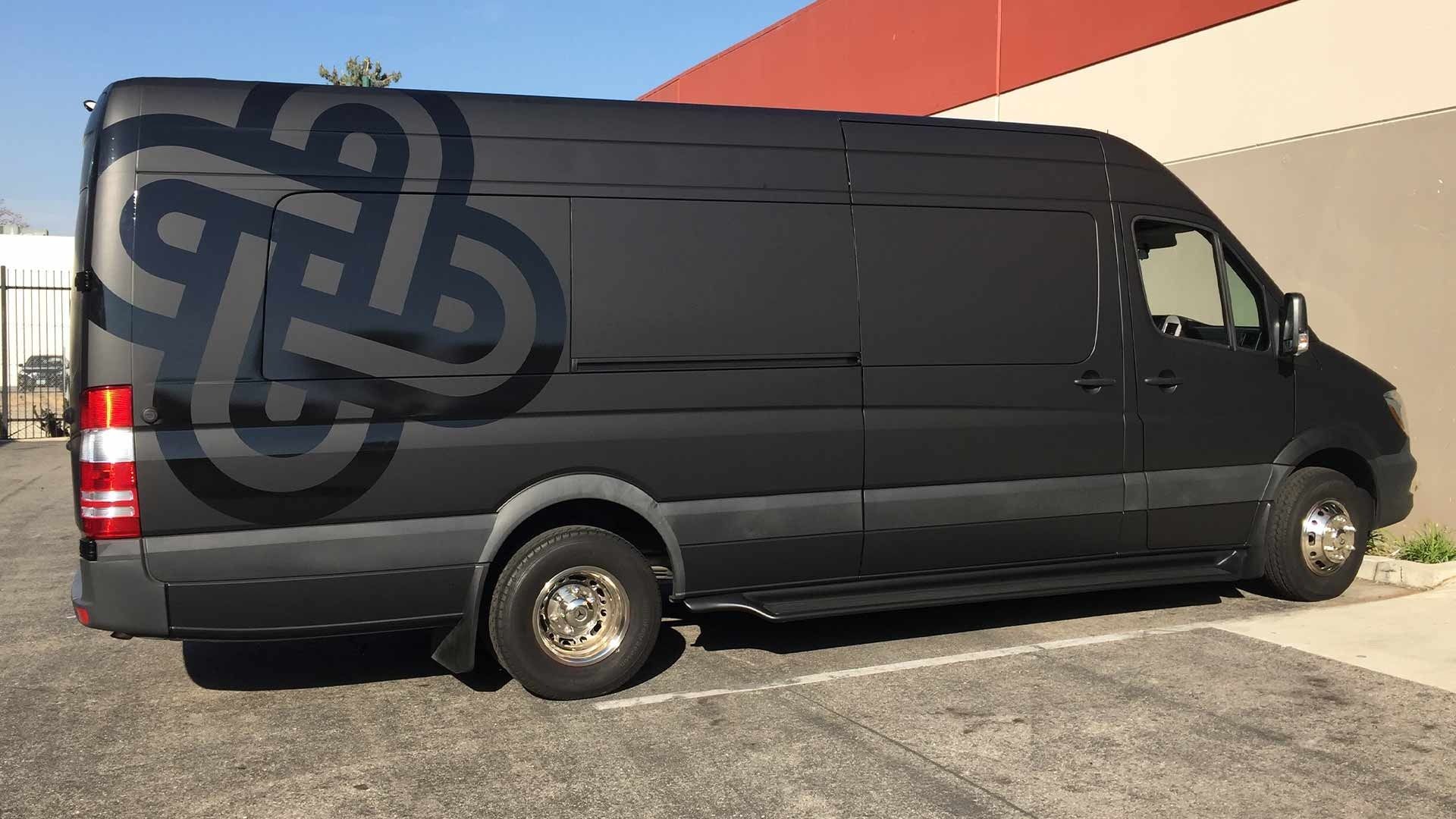 A matte black cargo van parked in a lot, featuring a large, glossy black abstract geometric logo on its side panel.